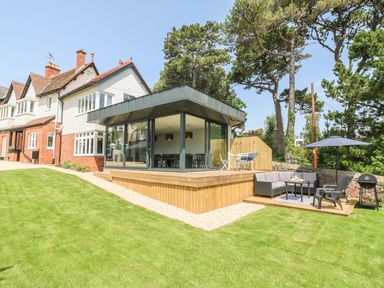 A modern house with a glass extension and wooden deck in a garden with patio furniture and a barbecue at The Coach House in Teignmouth