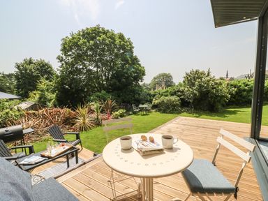 A wooden deck with a round table and two chairs overlooking a garden with trees and a grill at The Coach House in Teignmouth