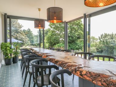 A dining room with a long wooden table surrounded by black chairs and large windows showing greenery at The Coach House in Teignmouth