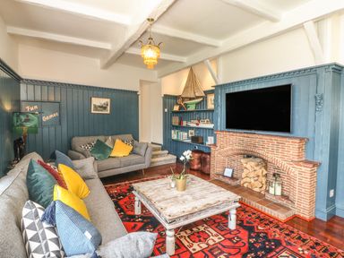 A living room with two sofas colorful pillows a wooden coffee table a red patterned rug a brick fireplace with stacked logs and a mounted TV at The Coach House in Teignmouth