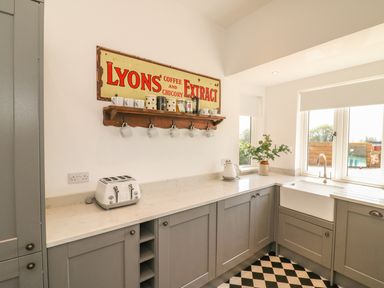 A kitchen with grey cabinets a white countertop a toaster kettle mugs on a wall shelf and windows above a sink at The Coach House in Teignmouth