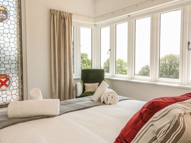 A bedroom with a bed, rolled towels, a green armchair, beige curtains, stained glass window, and multiple windows showing trees at The Coach House in Teignmouth