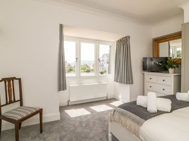 A bedroom with a bed with towels on it a chest of drawers with a TV and plant a striped chair and three windows with curtains at The Coach House in Teignmouth