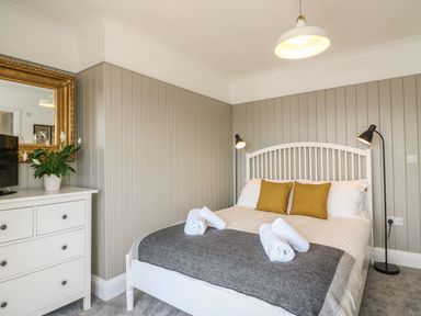 A bedroom with a white bed frame two mustard pillows rolled white towels on the bed gray blanket bedside lamps a white dresser with a plant and a gold mirror at The Coach House in Teignmouth