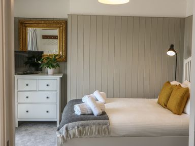 A bedroom with a bed having three rolled towels and two pillows a chest of drawers with a TV and a plant a large framed mirror and a floor lamp at The Coach House in Teignmouth
