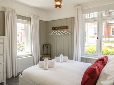 A bedroom with a bed with white linens and red pillows two windows with white curtains a wooden chair and a hanger rack on the wall at The Coach House in Teignmouth