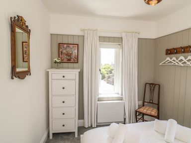 A bedroom with a white chest of drawers a window with curtains a wooden chair a wall mirror and white towels on a bed at The Coach House in Teignmouth