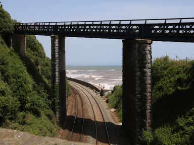 Railway tracks passing under a stone and metal bridge with the ocean and waves visible beyond the bridge