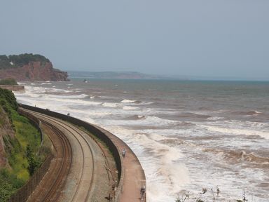 A coastal railway track next to the sea with waves crashing against the seawall and cliffs in the background at The Coach House in Teignmouth
