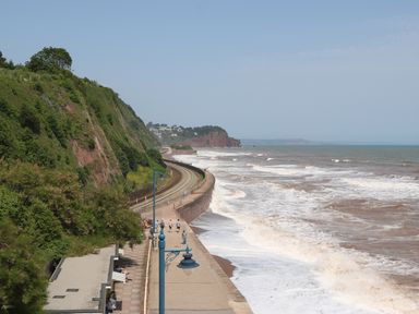 A coastal walkway with people walking next to railway tracks and a cliff with vegetation by the sea at The Coach House in Teignmouth
