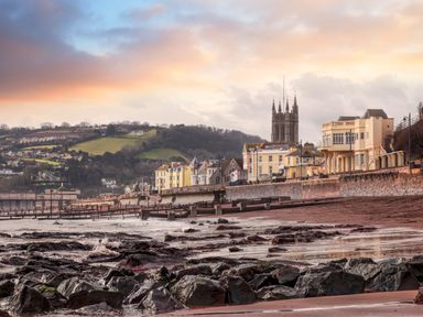 A rocky beach with waves and a row of buildings along a seawall with a church tower and hills in the background at The Coach House in Teignmouth