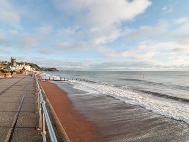 A sandy beach with waves and a paved promenade with a metal railing and buildings in the background at The Coach House in Teignmouth
