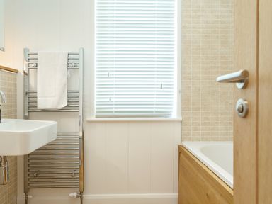 A bathroom with a towel on a metal towel rack a sink a window with blinds and a bathtub with a wooden panel at 3 Valley View in Lanreath