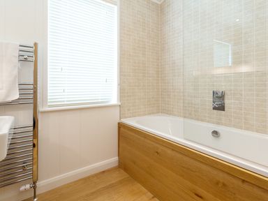 A bathroom with a bathtub with wooden paneling beige tiled wall a window with blinds and a towel rail at 3 Valley View in Lanreath