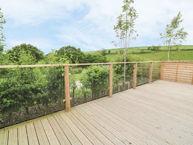 An outdoor wooden deck with glass and wood railing overlooking green fields and trees at 3 Valley View in Lanreath