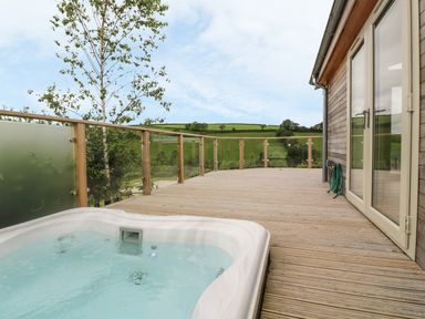 An outdoor wooden deck with a hot tub and glass railing overlooking green fields at 3 Valley View in Lanreath