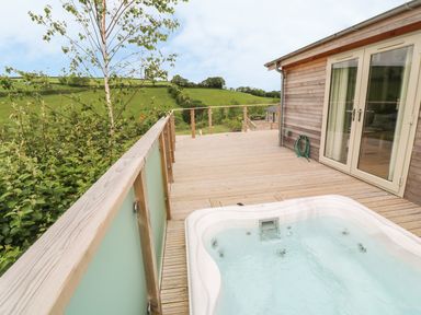 A wooden deck with a hot tub a garden hose and glass doors opening to a wooden house with green hills in the background at 3 Valley View in Lanreath