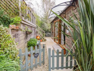 An outdoor walkway with a wooden fence and gate leading to a wooden building with an open door and plants along the sides at Wellinghill Lodge in Cheltenham