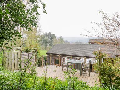 A wooden deck with a table and two chairs surrounded by greenery and a wooden fence at Wellinghill Lodge in Cheltenham