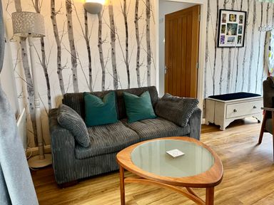 A living room with a grey sofa and teal cushions a wooden coffee table a floor lamp and framed photos on the wall at Wellinghill Lodge in Cheltenham