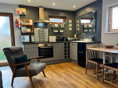 A kitchen with grey cabinets an oven a microwave a dining table with chairs and an armchair at Wellinghill Lodge in Cheltenham