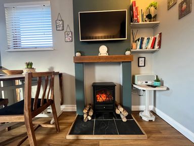 A living room with a fireplace under a wall-mounted TV a wooden dining table and chairs and shelves with books and plants at Wellinghill Lodge in Cheltenham