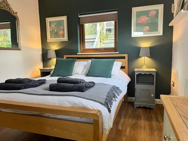 A bedroom with a wooden bed frame green pillows white bedding black towels bedside tables with lamps and framed floral pictures at Wellinghill Lodge in Cheltenham