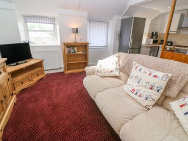 A living room with a beige sofa with patterned cushions a wooden TV stand with a television a wooden bookshelf a red carpet and an open kitchen area at The Stables in Longrock