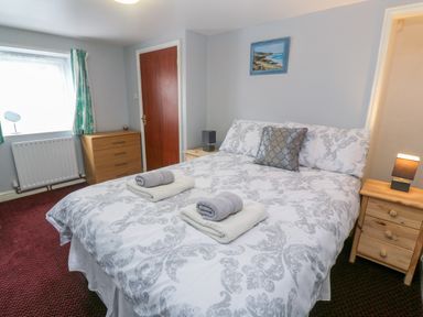 A bedroom with a double bed white and gray patterned bedding wooden nightstands with lamps a wooden chest of drawers and a window with green curtains at The Stables in Longrock
