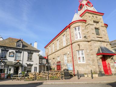 An outdoor seating area with wooden tables and benches next to a stone building with a clock tower and a white building with black trim at The Stables in Longrock