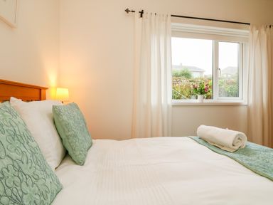 A bedroom with a made bed with green pillows and a rolled towel on the bed near a window with white curtains at Sunset in St Just