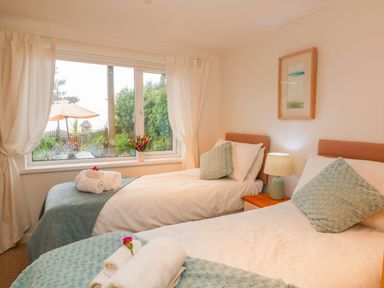 A bedroom with two single beds with white linens and blue throw pillows and blankets next to a window with curtains at Sunset in St Just