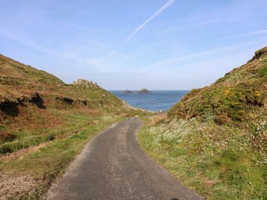 A paved road between grassy hills leading to the sea with small rocky islands visible on the horizon at Sunset in St Just