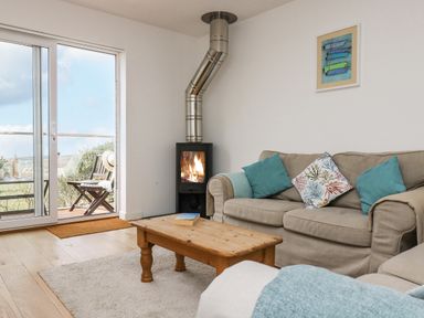 A living room with a beige sofa with blue and patterned cushions a wooden coffee table a fireplace and a sliding glass door leading to an outdoor seating area at Sunset in St Just
