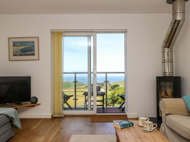 A living room with a sliding glass door opening to a balcony overlooking green hills and the sea at Sunset in St Just