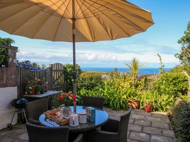 A patio with a round table and chairs under a large umbrella with plants and a view of the sea at Sunset in St Just