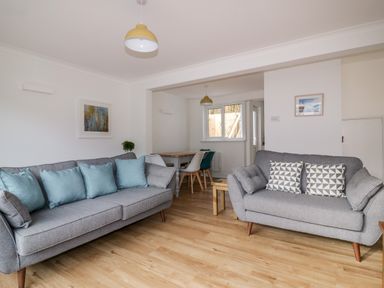 A living room with grey sofas with cushions a small wooden table and a dining table with chairs at Avalon in St Agnes