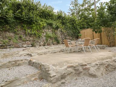 An outdoor patio area with a glass table and four wicker chairs surrounded by stone walls and gravel at Avalon in St Agnes