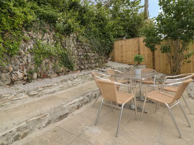 An outdoor patio with a metal table and four wicker chairs next to a stone wall and wooden fence at Avalon in St Agnes