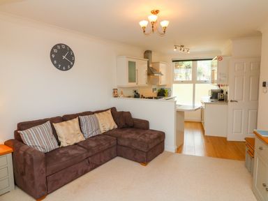 An open plan living room with a brown sofa with cushions a wall clock and a kitchen area at Seashore in Woolacombe