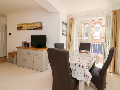 A dining area with a table covered by a tablecloth and four chairs next to a cabinet with a television at Seashore in Woolacombe