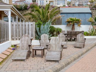 An outdoor patio with wooden lounge chairs and a small table near plants and a blue shed at Seashore in Woolacombe