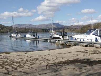 A dock with boats moored on a lake with hills and trees in the background at St Marys Cottage in Windermere