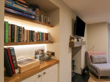 A room with wooden shelves holding books and board games a TV mounted above a fireplace and a gray chair with a pink blanket at St Marys Cottage in Windermere