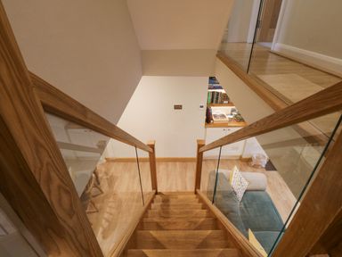 A wooden staircase with glass panels leading down to a living area with a green sofa and bookshelf at St Mary's Cottage in Windermere