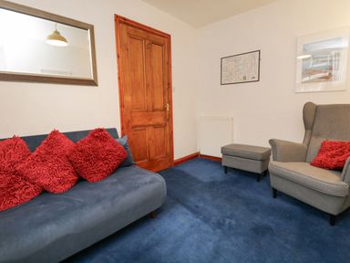 A room with a blue sofa with red cushions a gray armchair with a red cushion a footstool a wooden door a wall mirror and framed pictures at St Mary's Cottage in Windermere