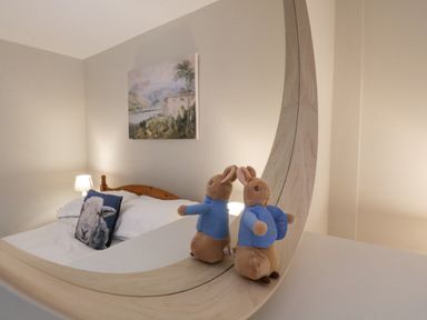 A bedroom with a bed that has white bedding and a sheep pillow with a wooden headboard a lamp on the bedside table and a painting on the wall with a plush rabbit toy reflected in a curved mirror at St Mary's Cottage in Windermere