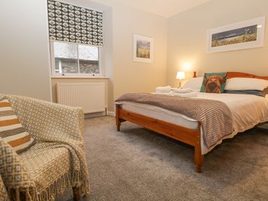 A bedroom with a wooden bed with white bedding and a dog pillow a patterned armchair and a window with a patterned blind at St Marys Cottage in Windermere