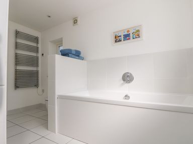 A bathroom with a white bathtub blue towels on a ledge a wall-mounted towel heater and a framed picture on the wall at The Upper Deck in Porthcurno