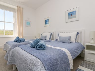 A bedroom with two single beds with blue bedding and rolled towels a window and framed pictures on the wall at The Upper Deck in Porthcurno